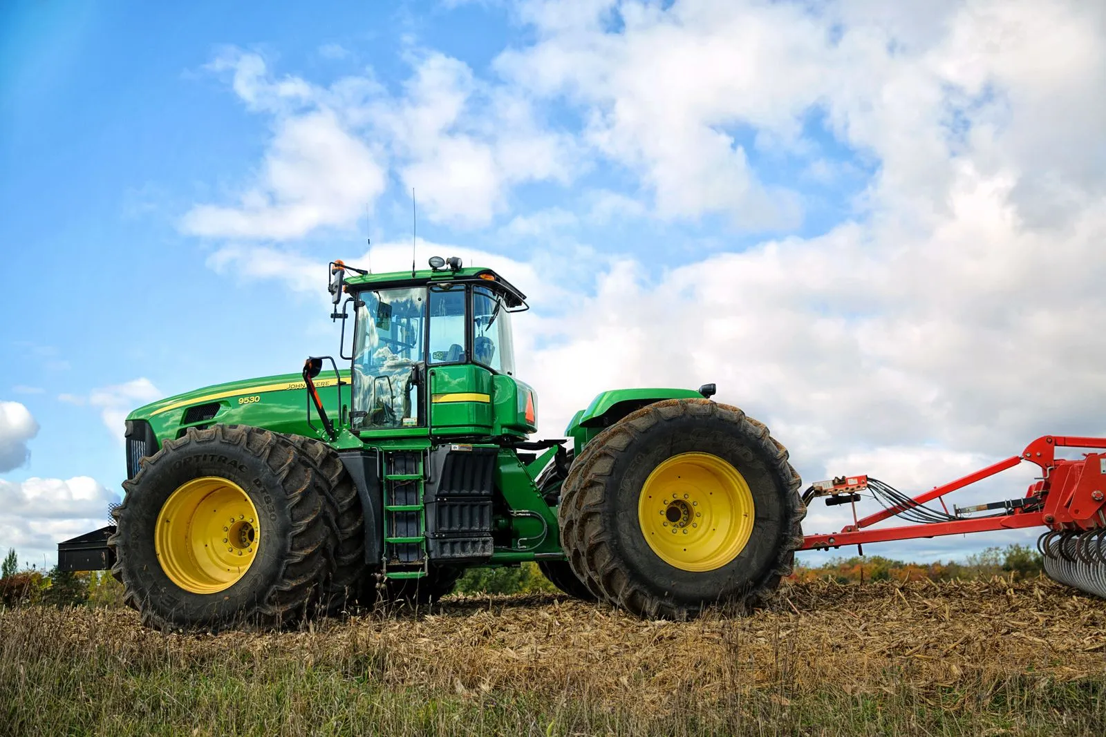 Green John Deere tractor pulling a red implement across a harvested field under a cloudy sky