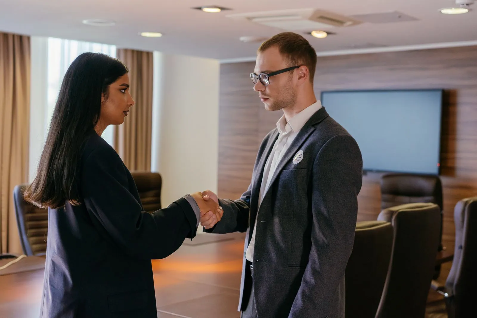 Man and woman shaking hands in a boardroom with a wall-mounted screen and empty chairs