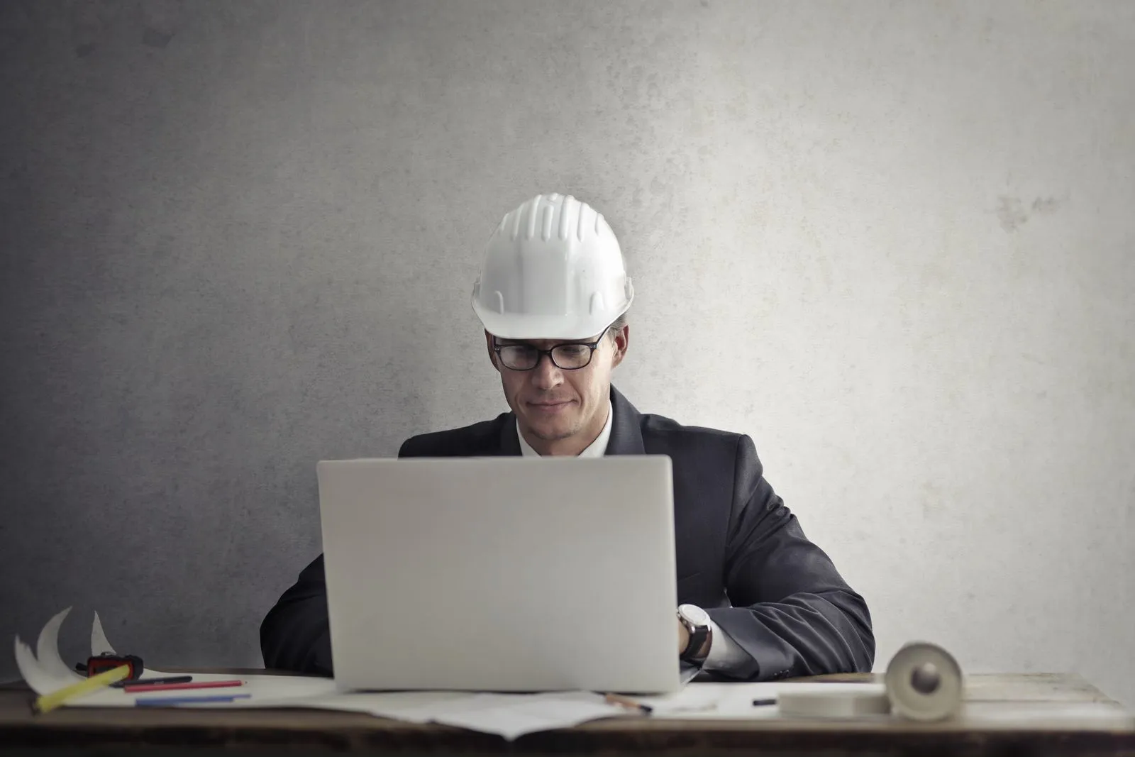 Man in a suit and white hard hat typing on a laptop next to construction tools on a desk