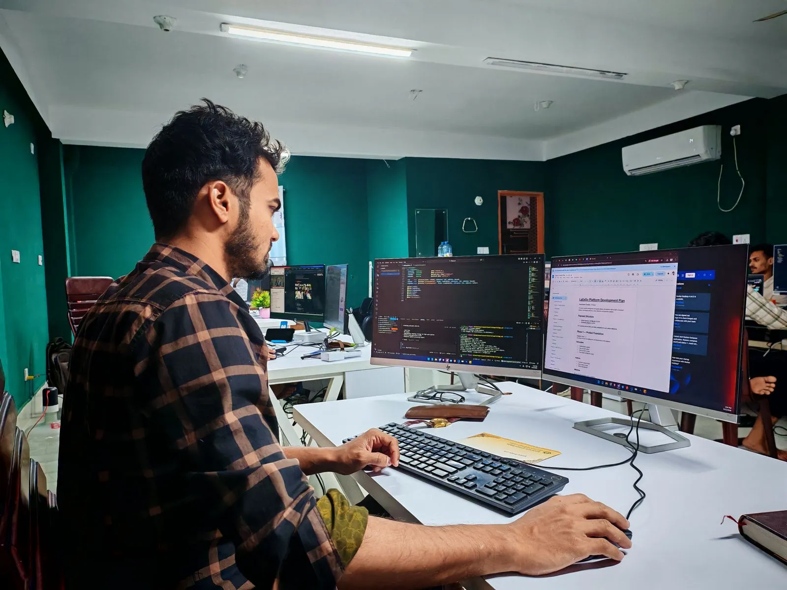Man viewing a code editor and a document across dual monitors in a teal-walled office