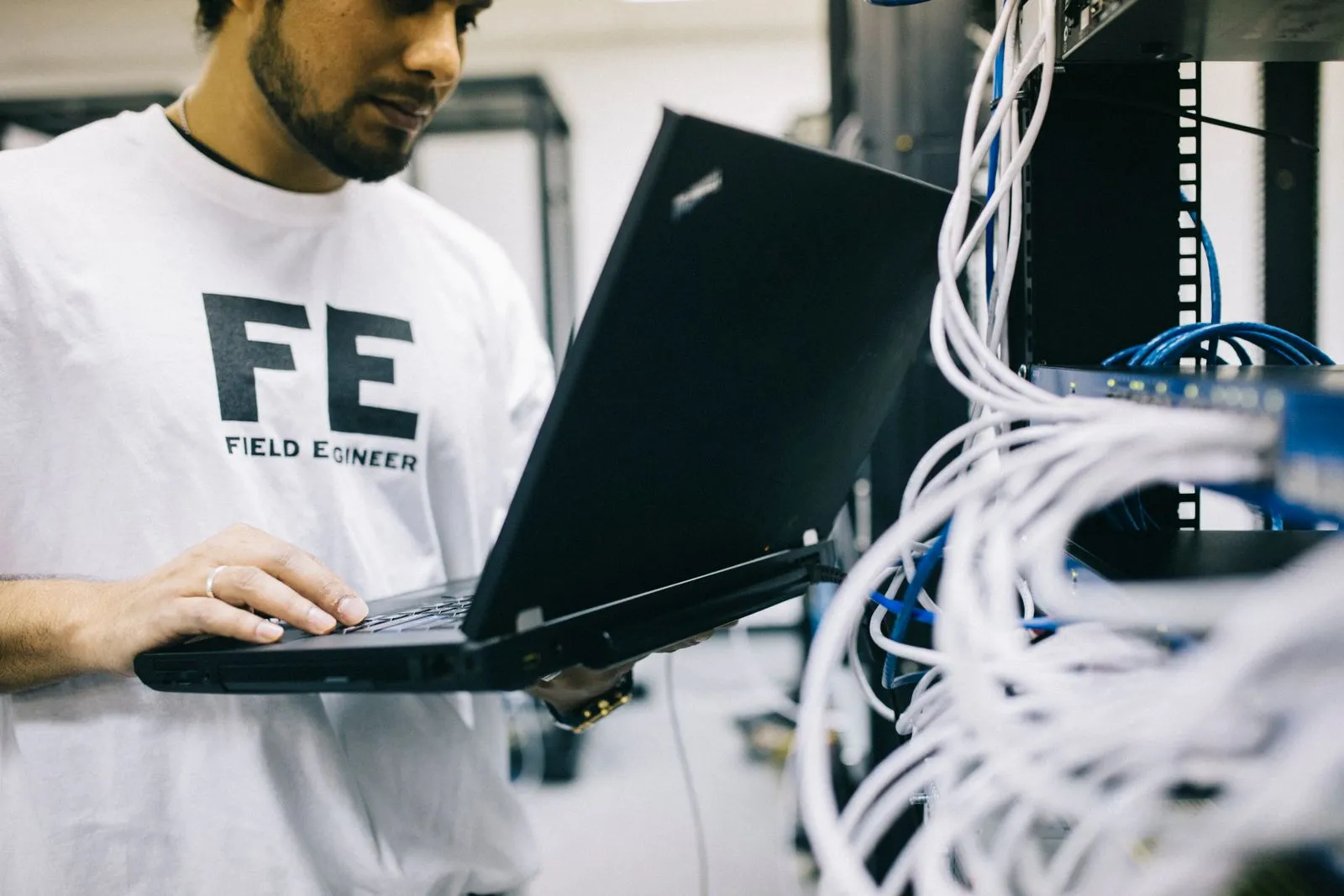 Man in a Field Engineer t-shirt holding an open laptop beside a server rack