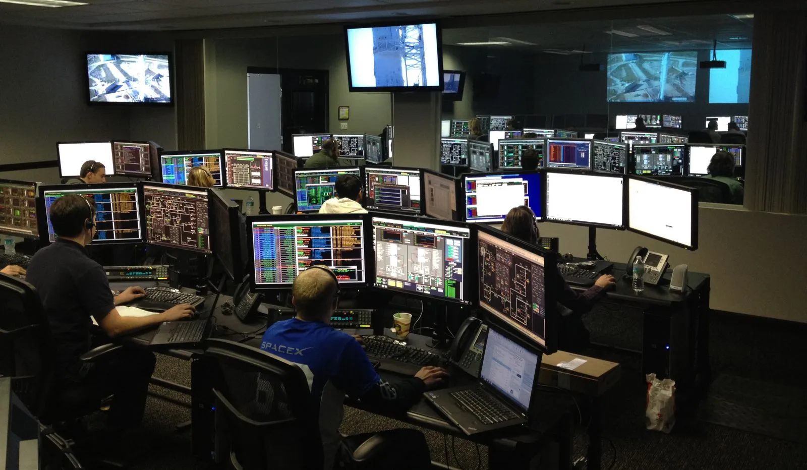 Mission-control room with staff at multi-monitor desks, one wearing a SpaceX shirt
