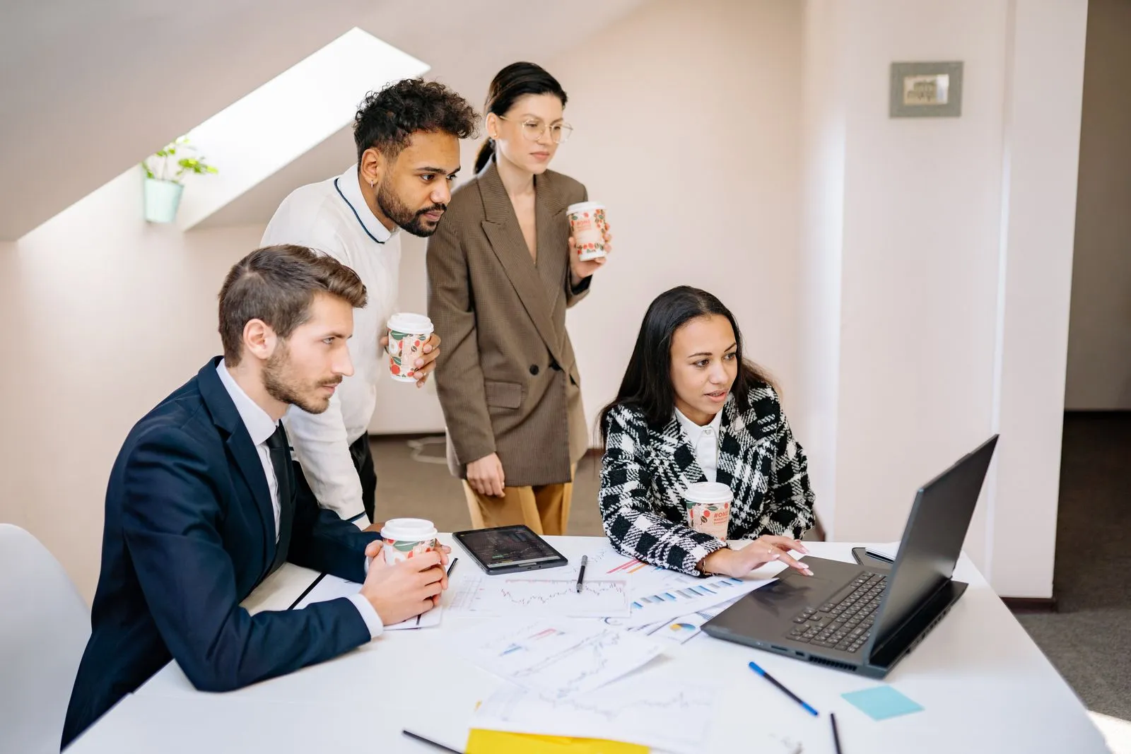 Four coworkers holding coffee gather around a laptop and printed charts on a desk