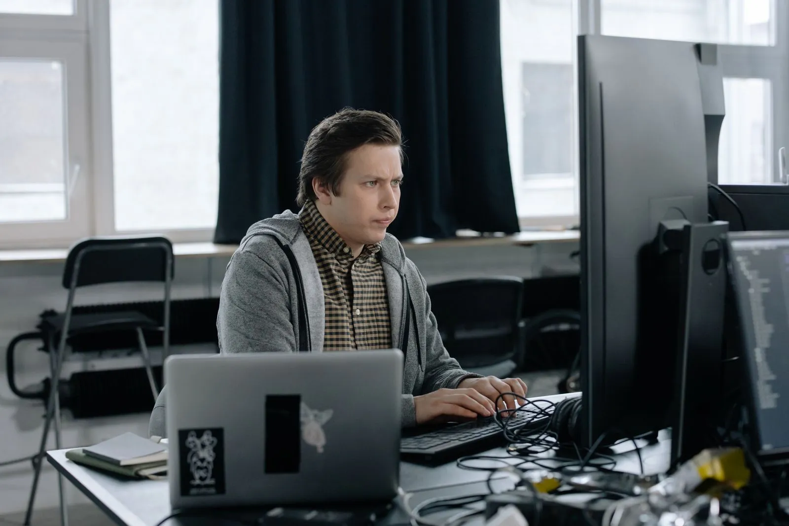 Man with a puzzled expression working at a laptop and dual monitors near curtained windows
