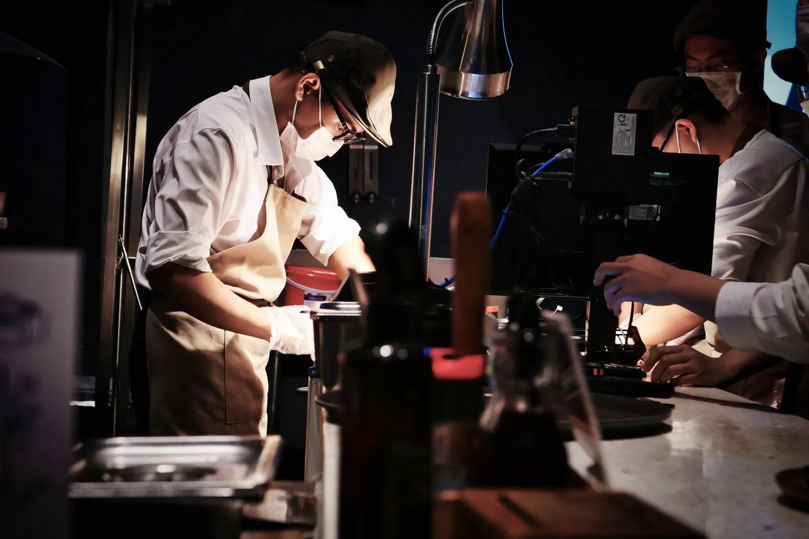 Workers in flat caps and face masks handle dark equipment under a work lamp at a counter