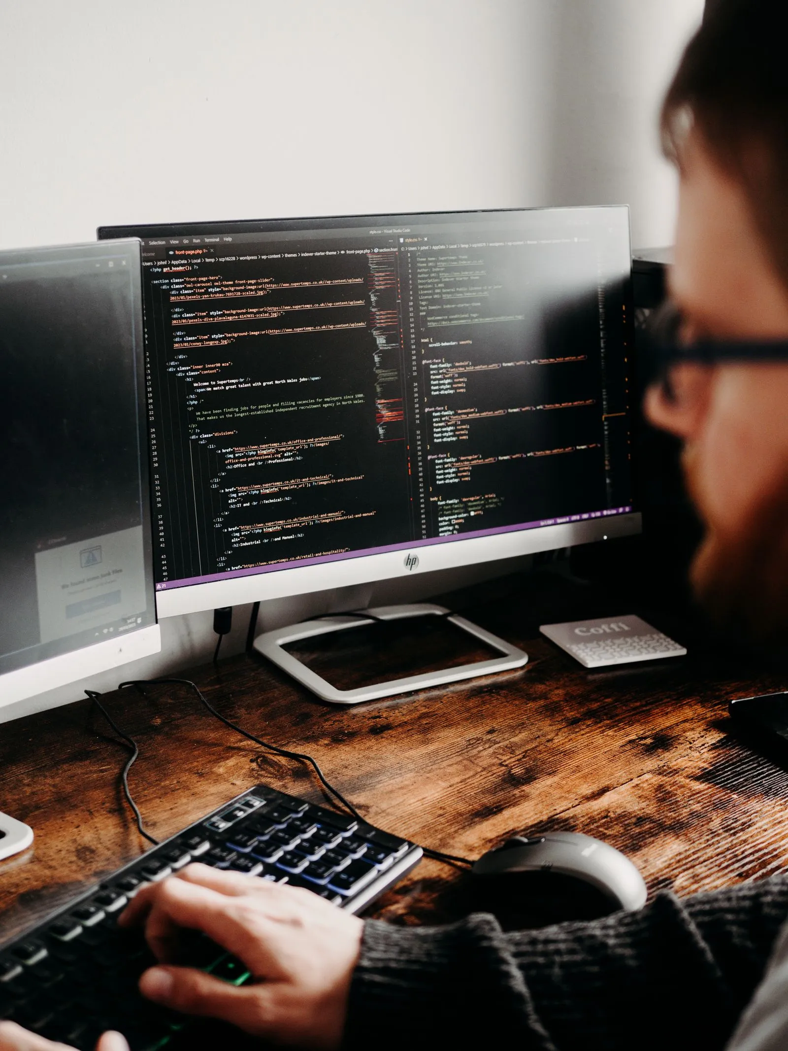 Man viewing PHP and CSS code for a WordPress theme across two monitors on a wooden desk