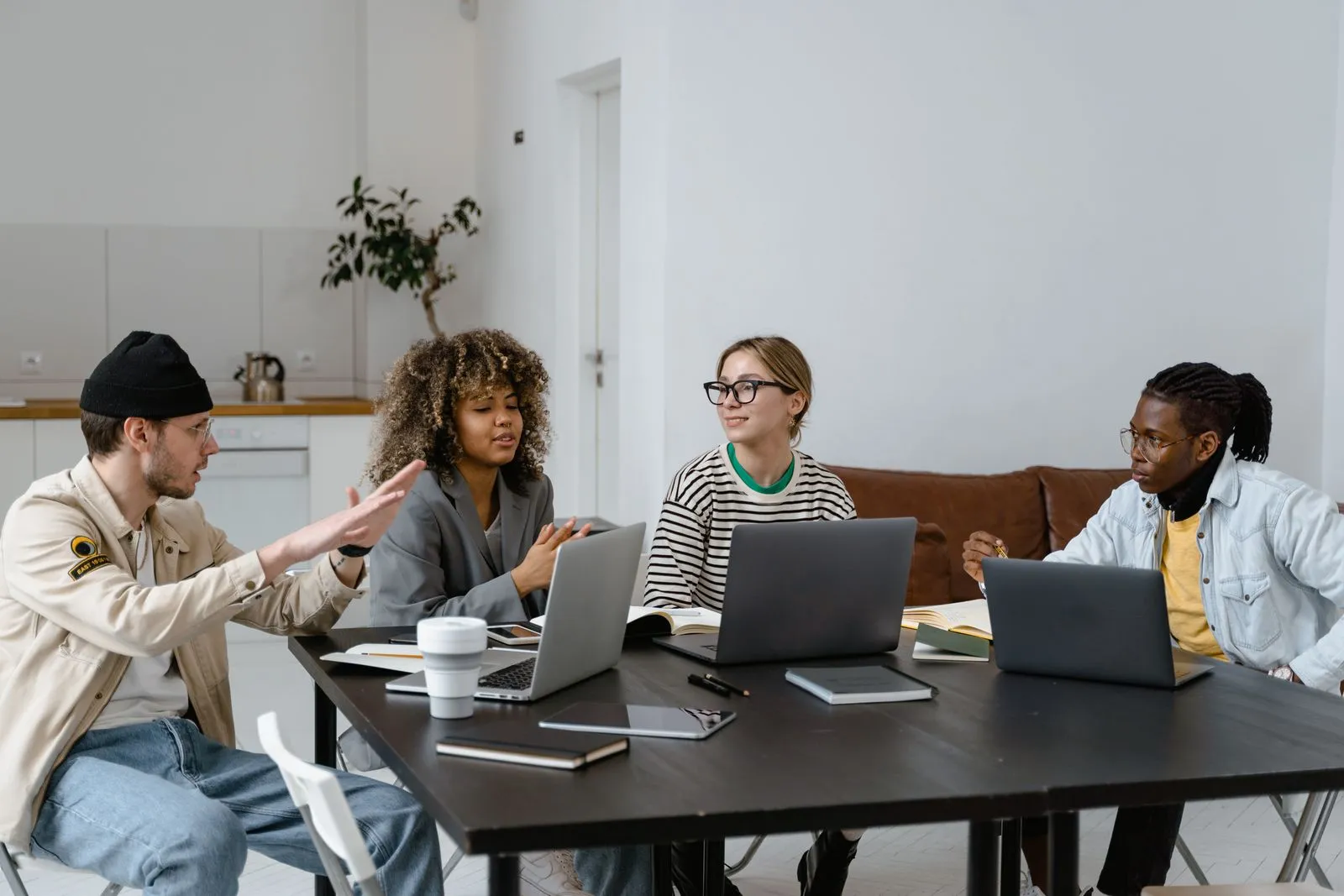 Four coworkers with laptops and notebooks talk around a table in a loft-style space with a kitchen behind
