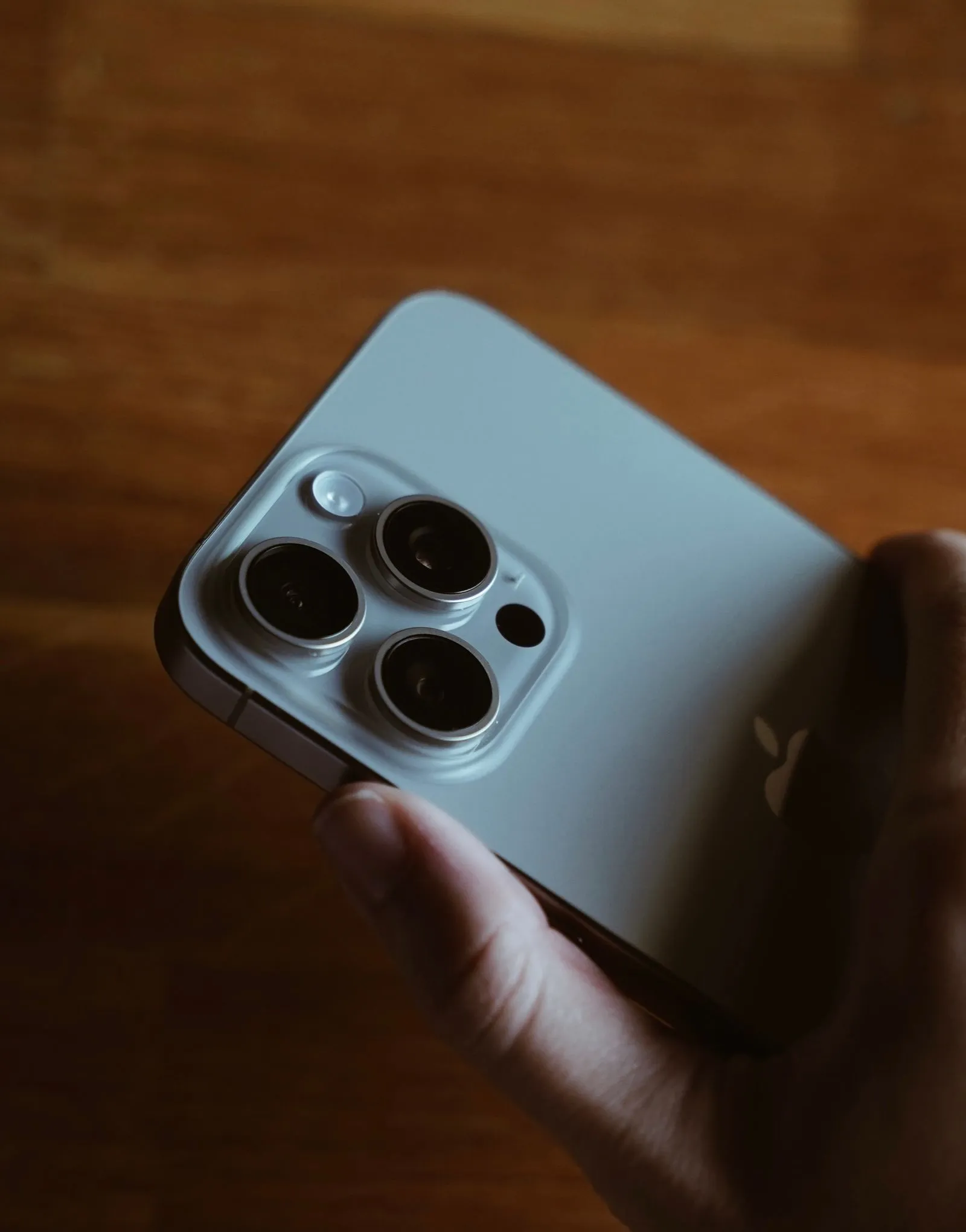 Hand holding a sage-green iPhone showing its triple-camera lens module on a wooden table