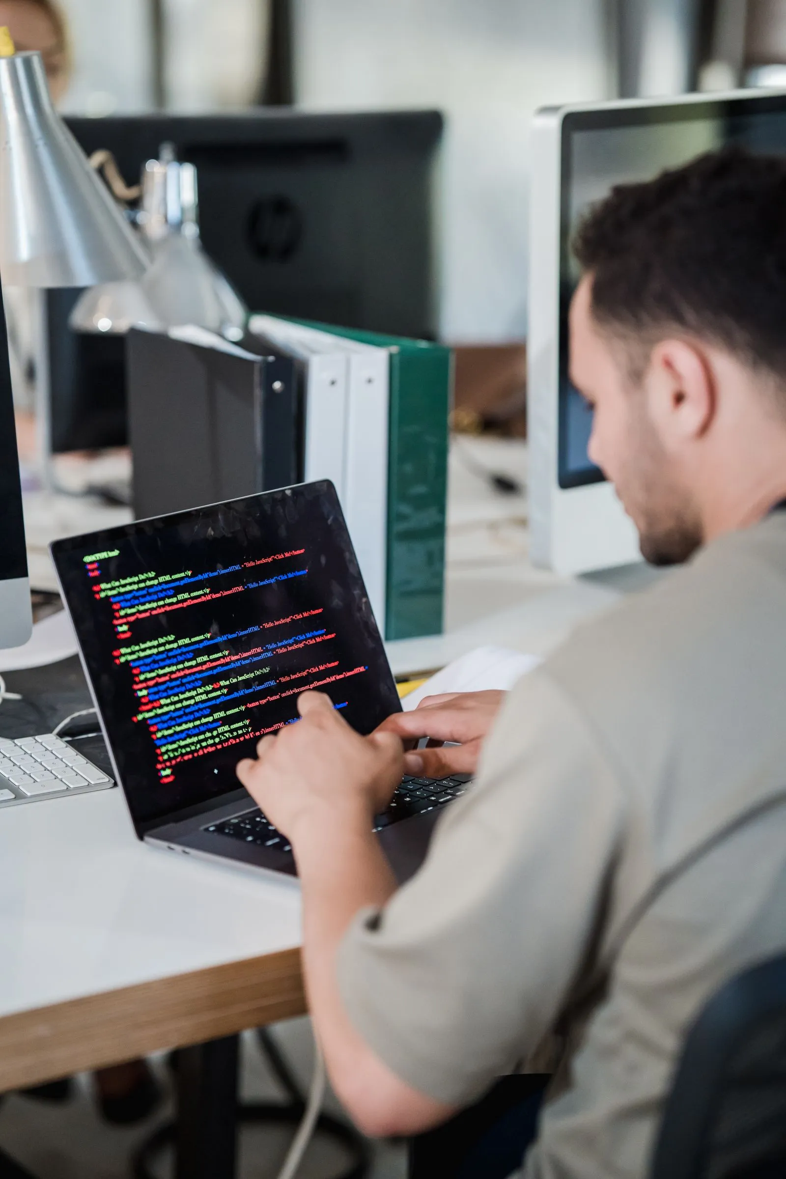 Man typing on a MacBook displaying colorful JavaScript code in an open-plan office