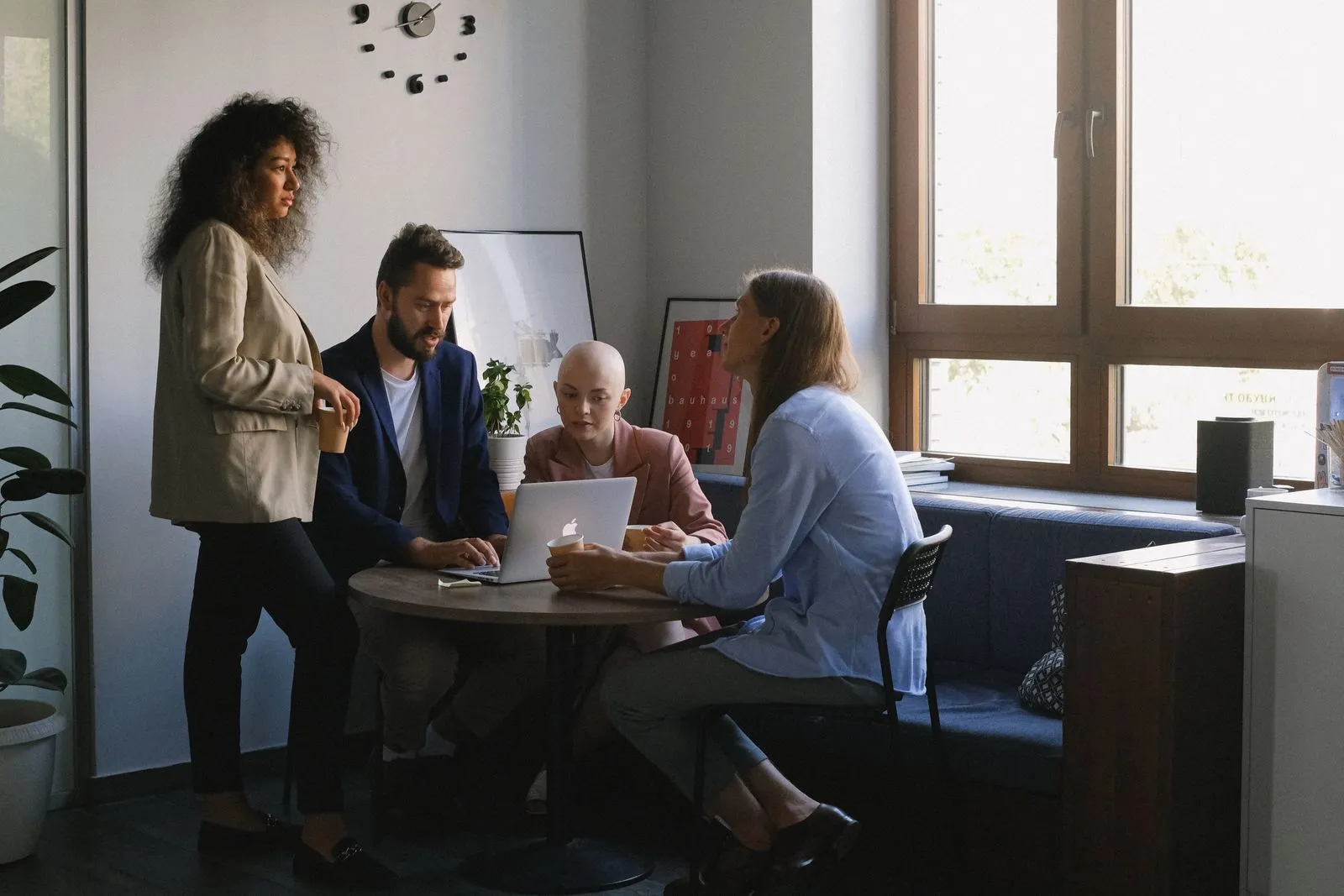 Four coworkers gathered around a laptop at a small table below a wall clock