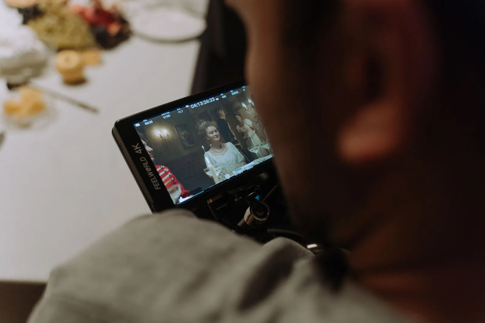 Camera monitor showing a costume drama scene on a film set, with a catering table blurred behind