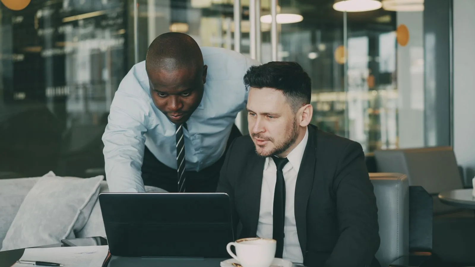 Two businessmen in suits reviewing a tablet together at a cafe table with coffee