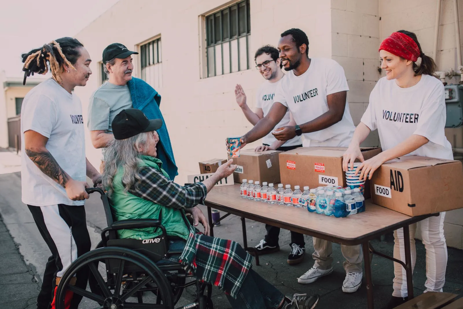 Volunteers in 'VOLUNTEER' t-shirts hand water and aid supplies to an elderly man in a wheelchair