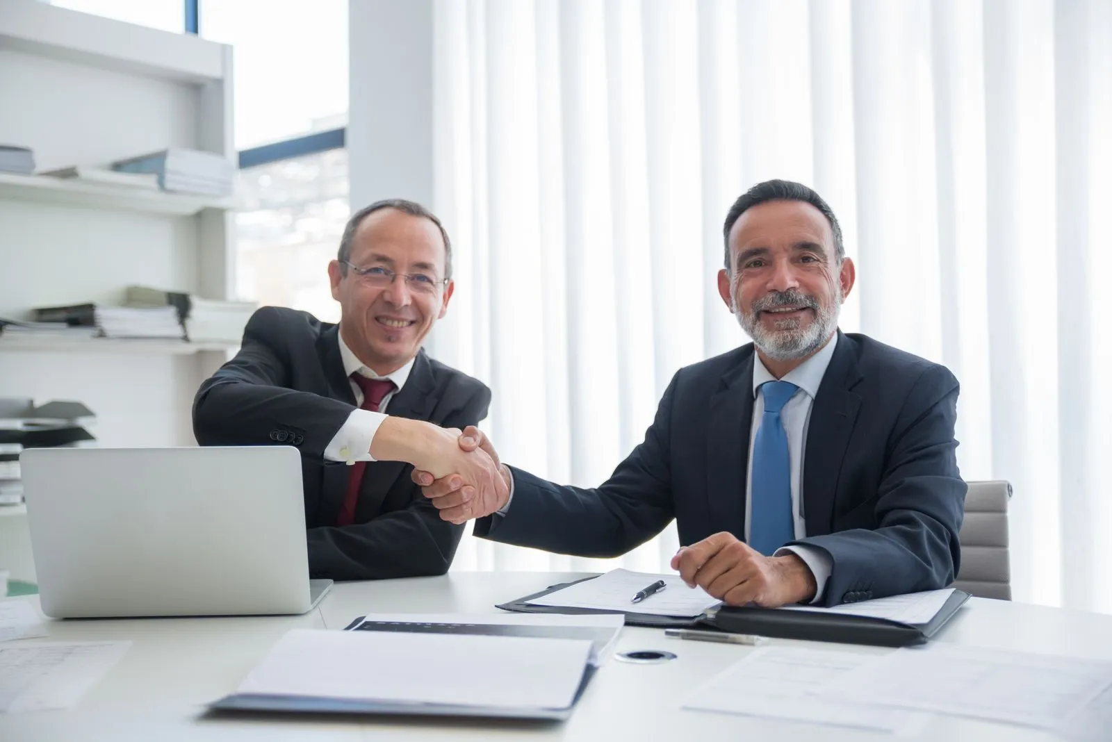 Two smiling businessmen in suits shake hands over an office desk with an open laptop