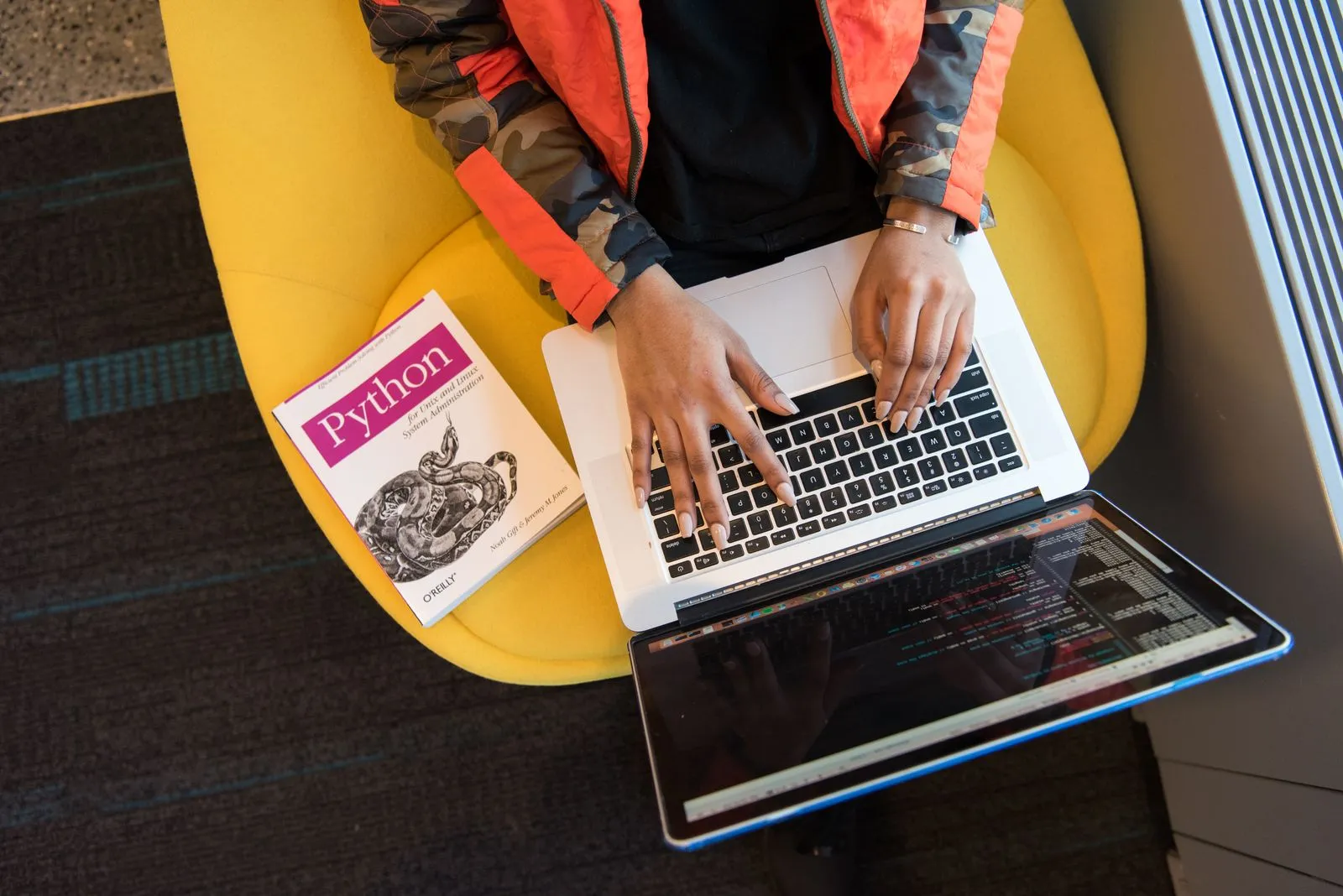 Overhead view of hands typing on a MacBook beside an O'Reilly Python book on a yellow chair