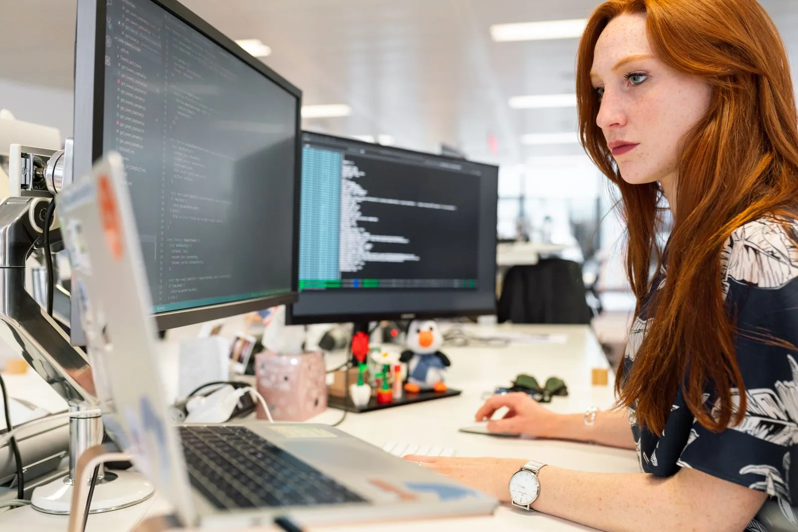 Red-haired woman viewing code on dual monitors, with a penguin plush toy on the desk
