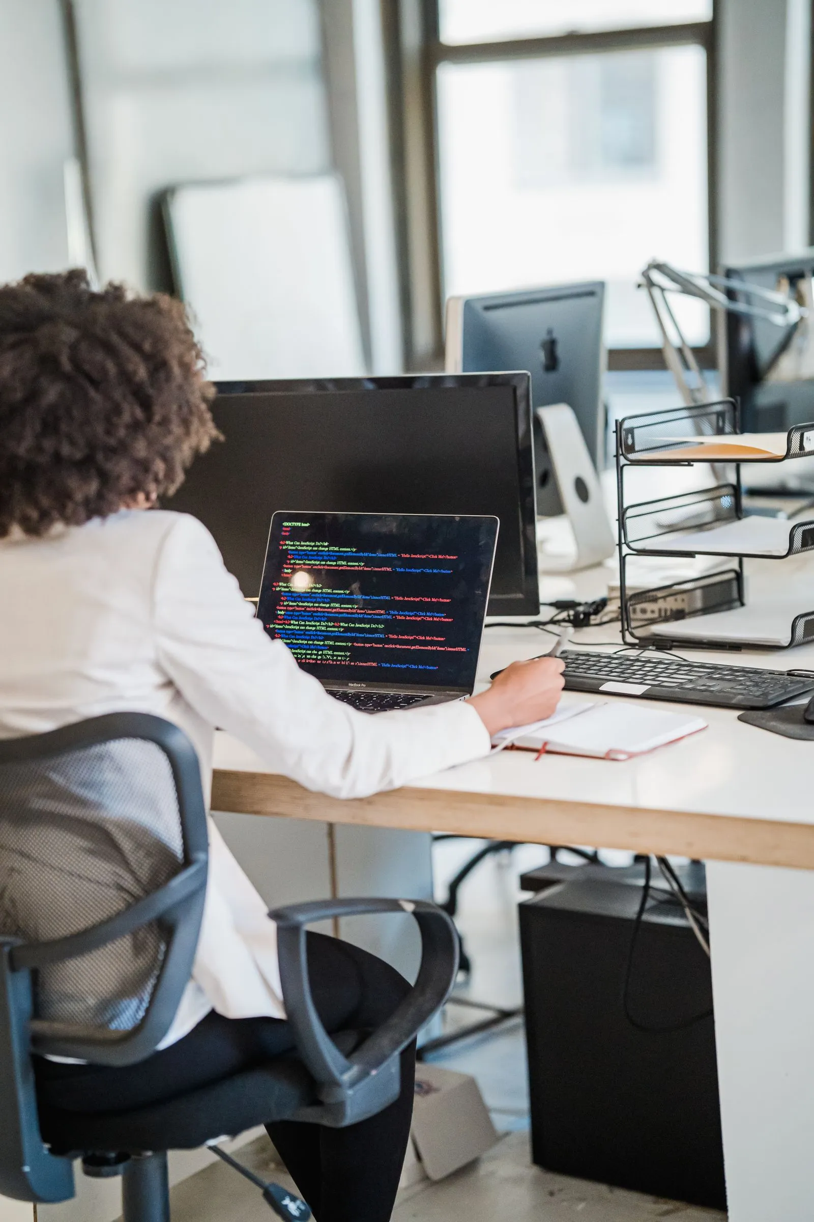 Woman with curly hair viewing JavaScript code on a laptop while holding a notebook