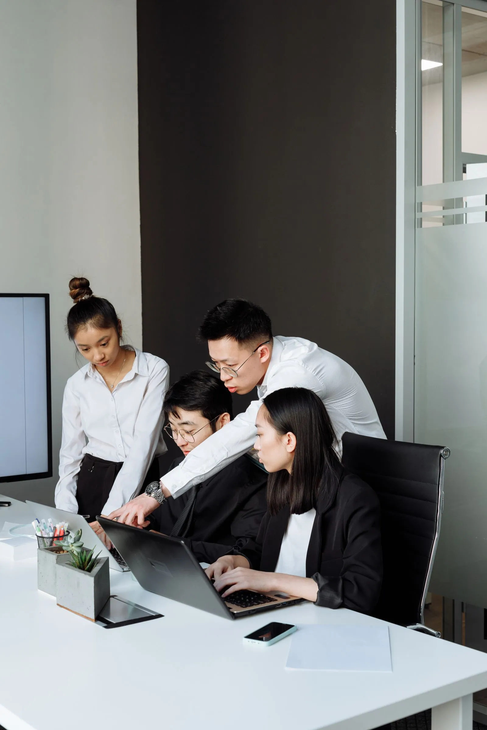 Four colleagues in a dark-walled office huddled around two laptops on a desk