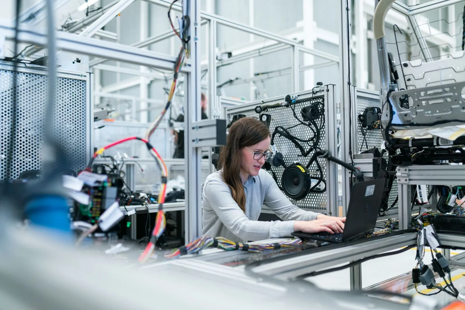 Woman typing on a laptop amid wiring harnesses and equipment racks in an industrial workshop