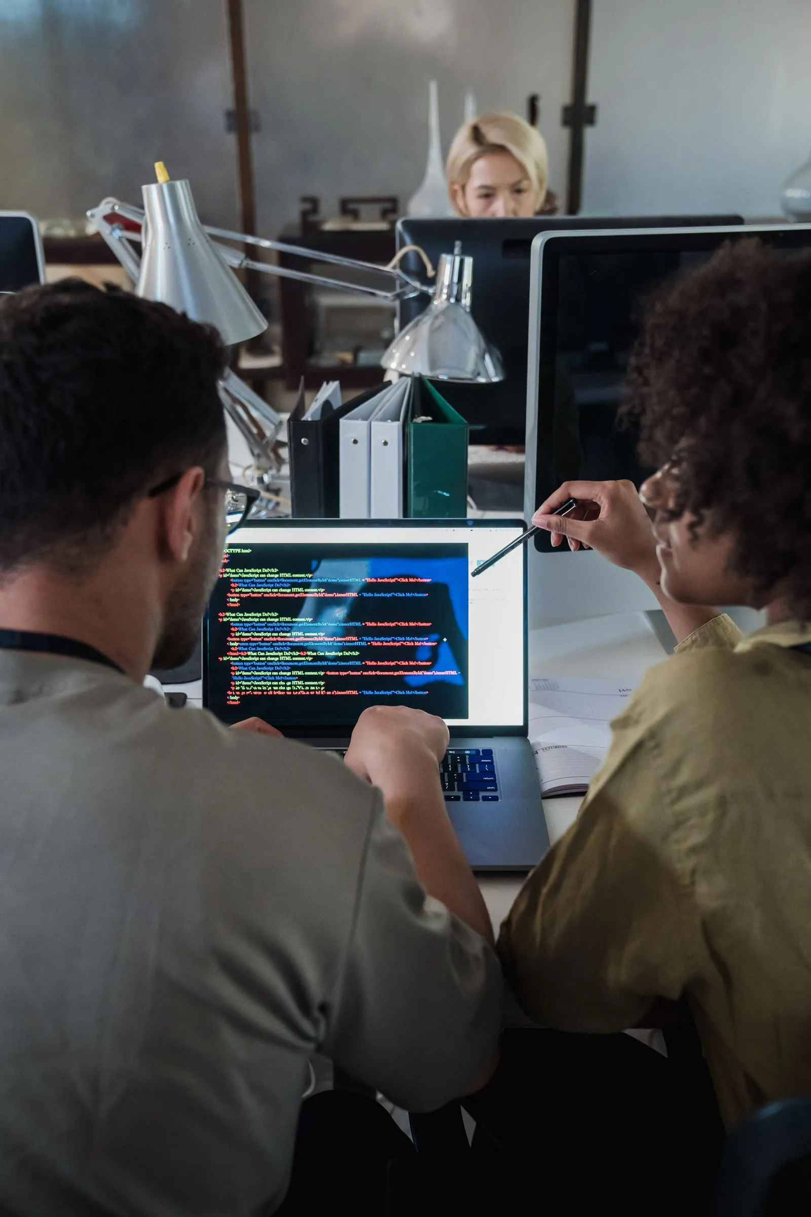 Two coworkers pointing at HTML and JavaScript code on a laptop screen at a shared desk
