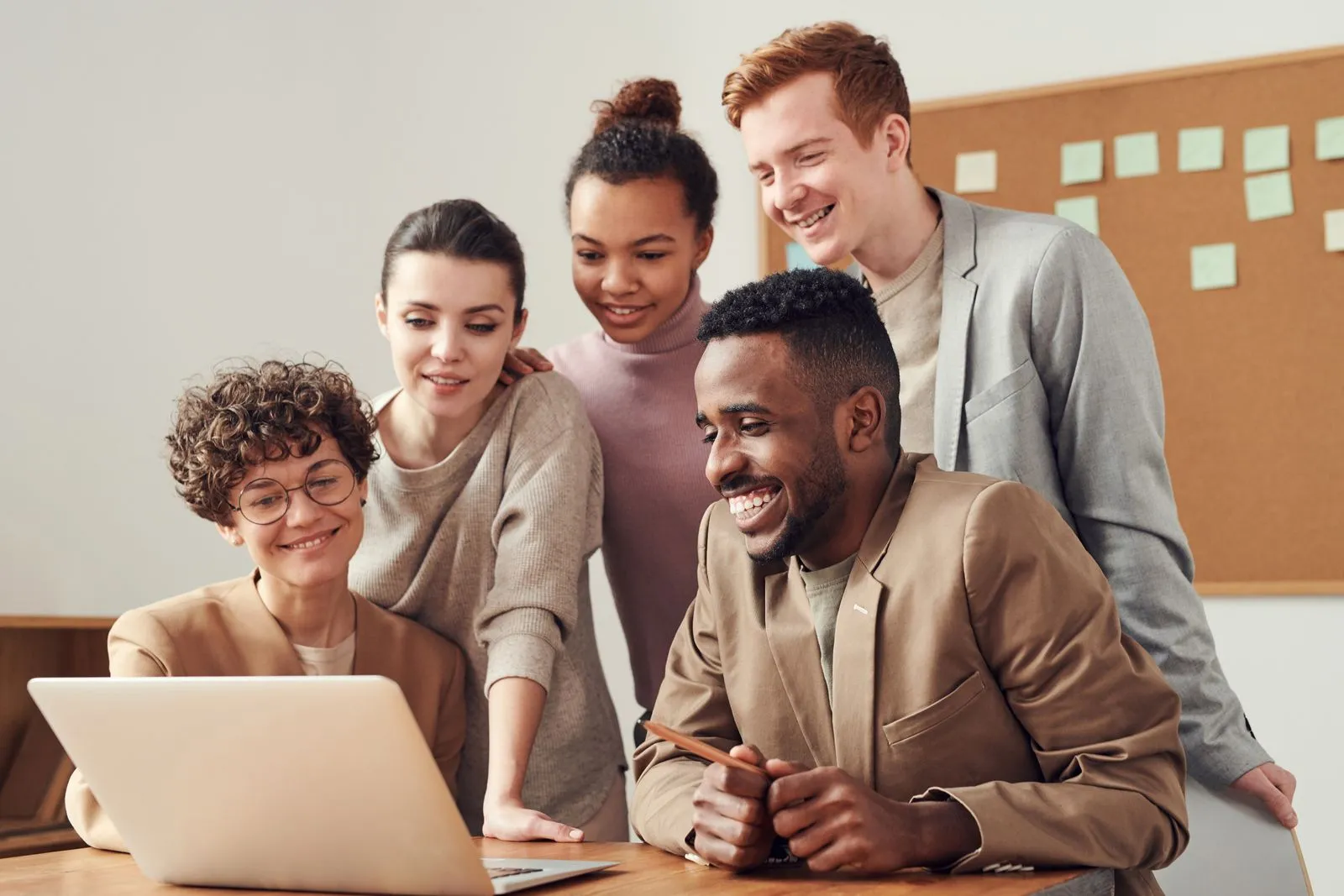 Five smiling coworkers gathered around a laptop in front of a cork board with sticky notes