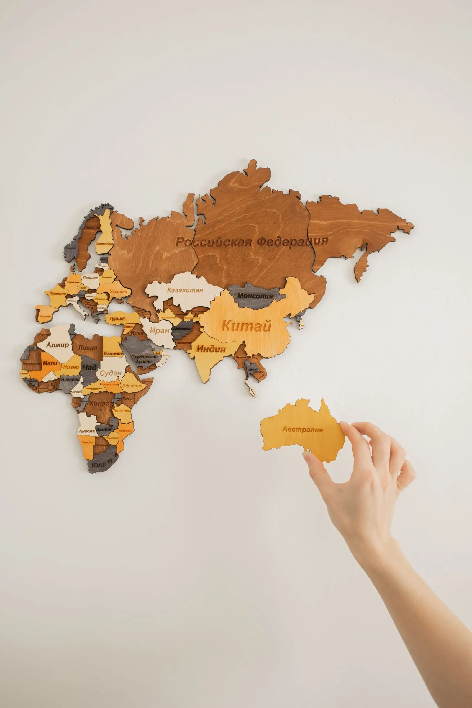Hand placing an Australia piece onto a wooden world map wall decoration labeled in Russian