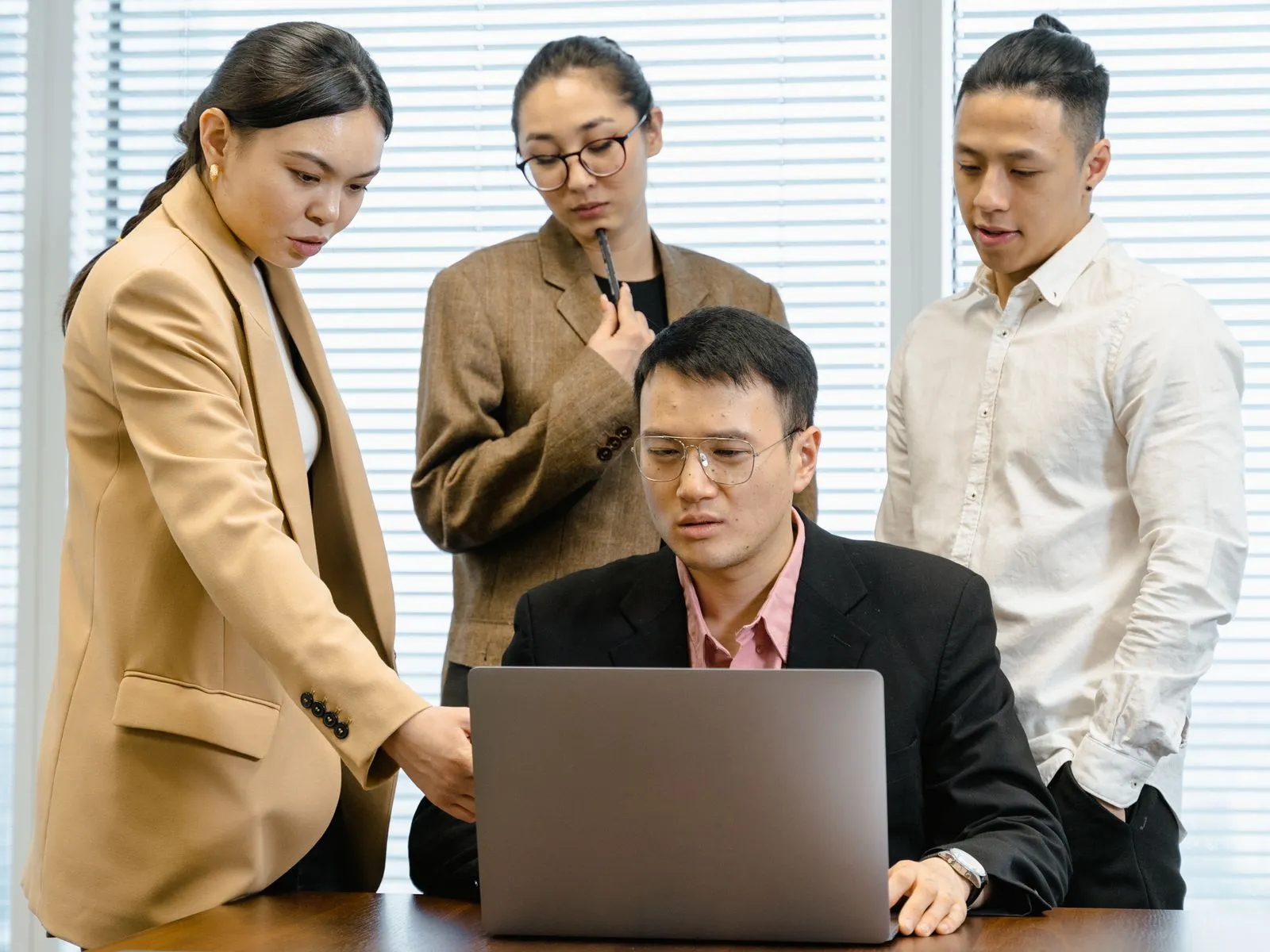 Three coworkers standing behind a seated colleague, all looking at a laptop screen in a blinds-lit office