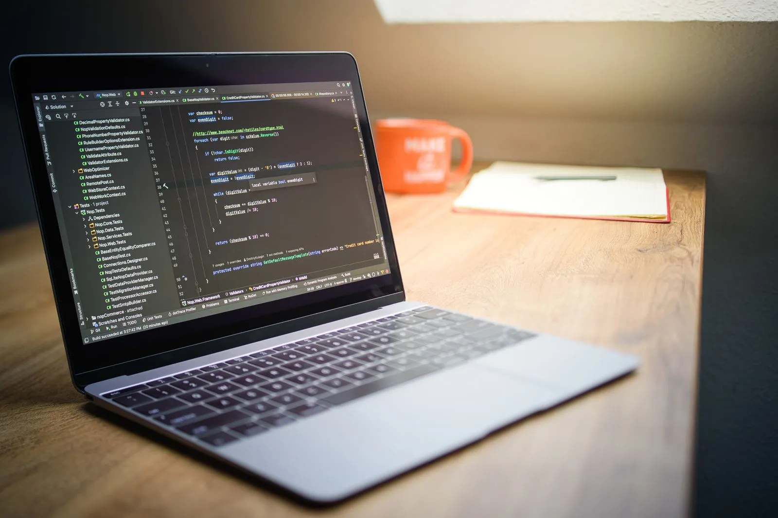 Laptop on a wood desk showing C# code in an IDE, beside an orange mug reading 'Make it Happen' and a notebook